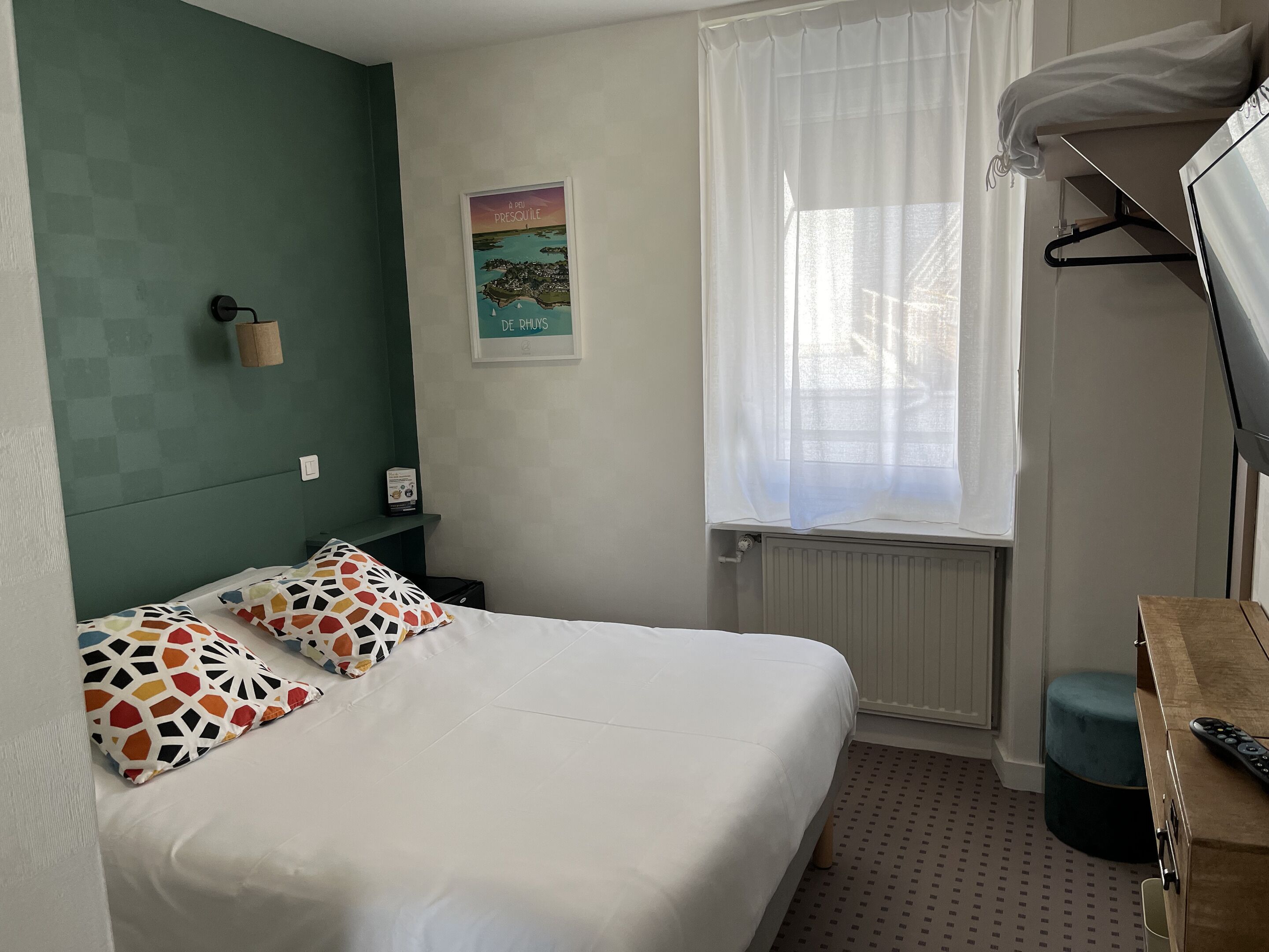 A neatly arranged hotel room with a bed, a window with sheer curtains, a small table with a remote control, and a wall-mounted shelf with a potted plant.