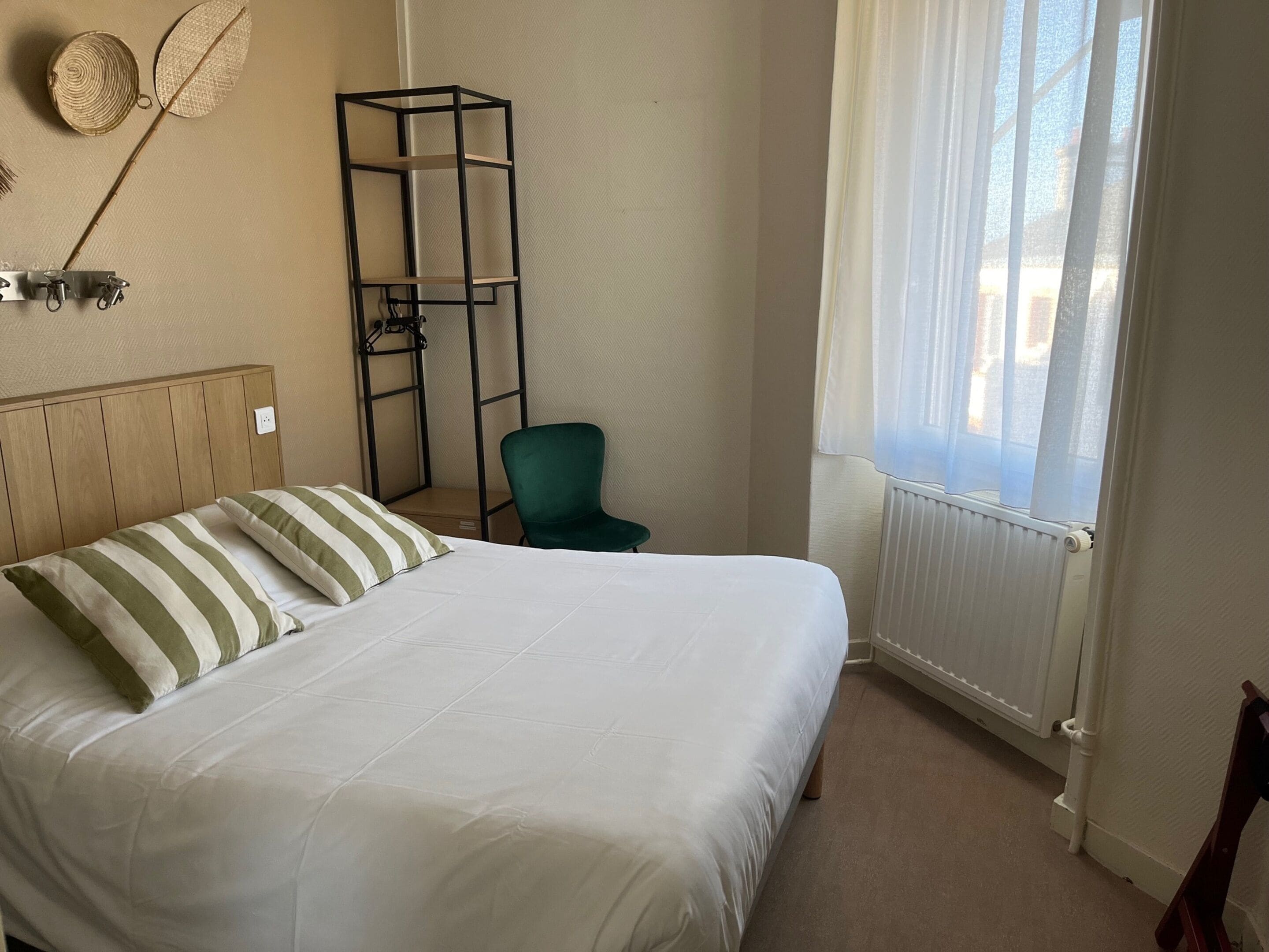 Una cama bien hecha con almohadones a rayas, una silla verde, un estante en la pared y una ventana con cortinas de gasa.