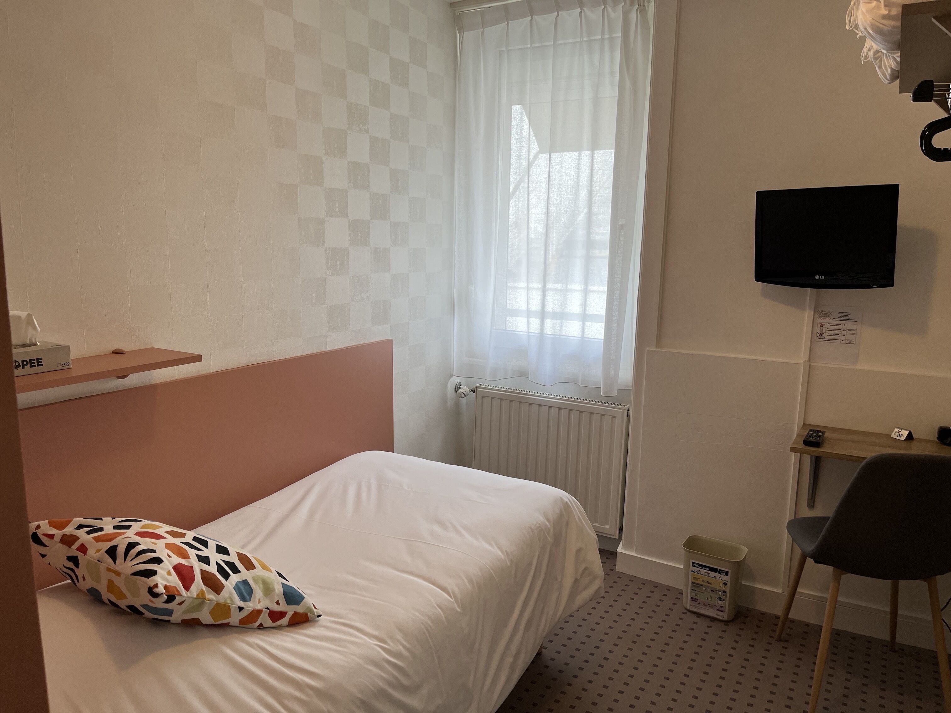 A hotel room with a bed, a desk, a chair, a TV, a radiator, and a window with curtains.