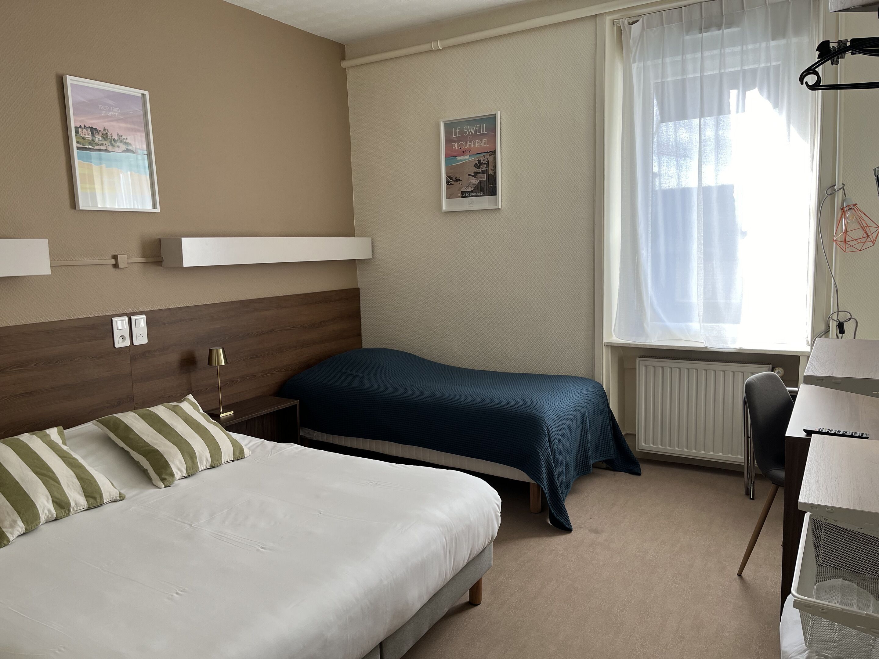 A hotel room with two beds, a desk, and a window with sheer curtains.