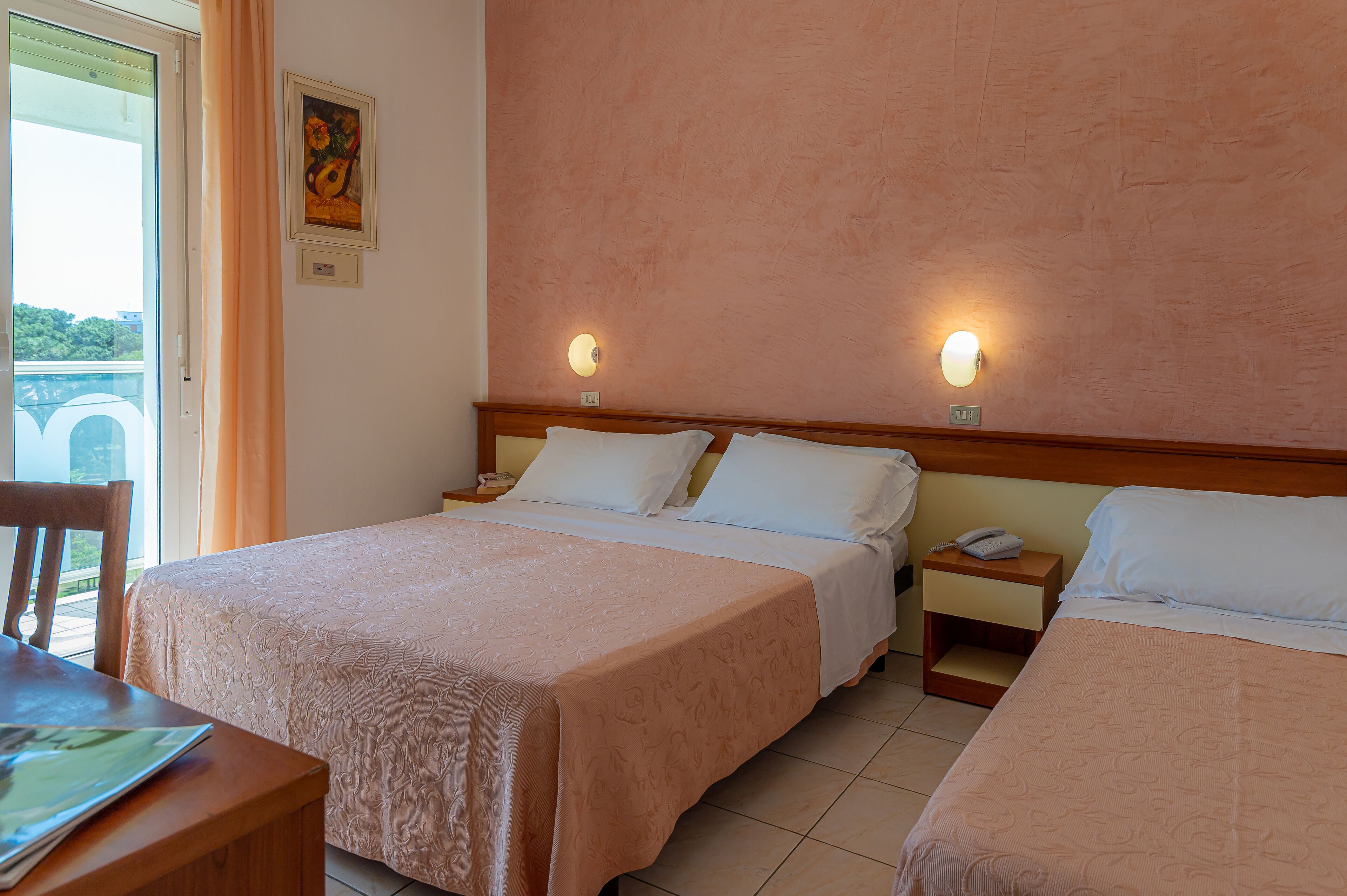 A hotel room with two beds, a wooden headboard, bedside tables with lamps, a chair, and a view of greenery outside.