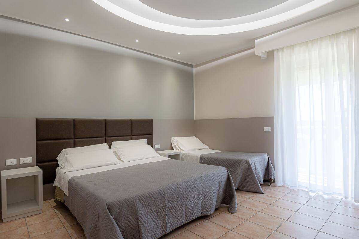 A modern hotel room with two beds, a built-in headboard, a bedside table, and a large window with sheer curtains.