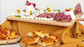 Uma tábua de madeira com diversos queijos, charcutaria e um queijo inteiro, acompanhada de pão fatiado e uma pizza.