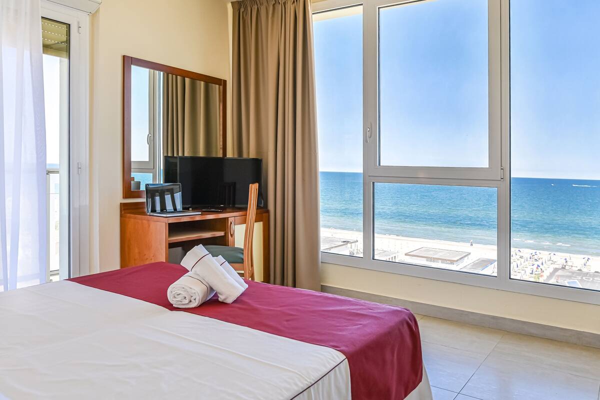 A hotel room with a bed, a desk with a TV, and a large window overlooking the beach.