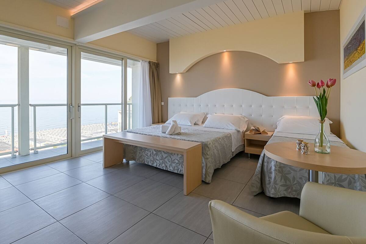 A hotel room with a large bed, a bedside table, a chair, and a view of the sea.