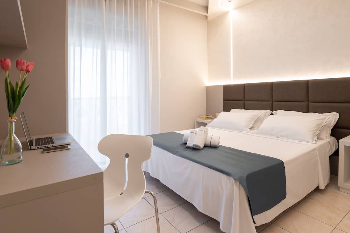 A modern hotel room with a neatly made bed, a desk with a laptop, a chair, and a vase with flowers.