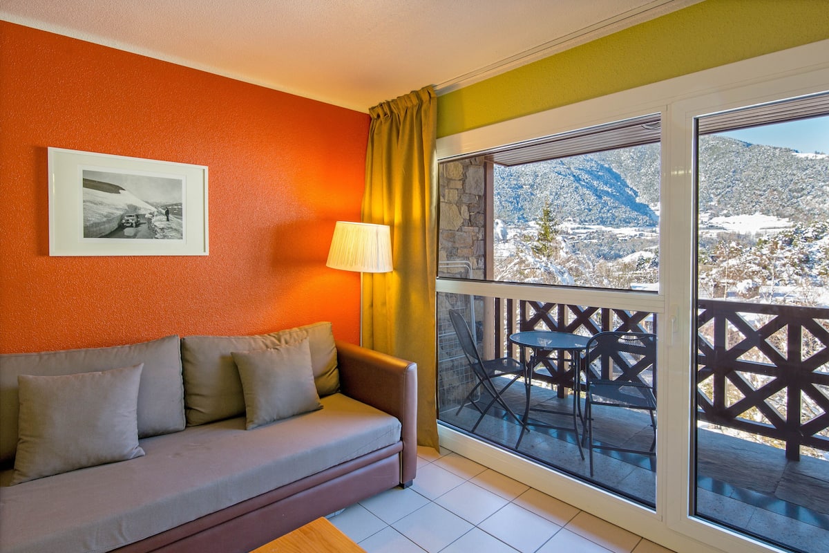 Quarto com sofá, quadro na parede, mesa com cadeiras e varanda com vista para montanhas cobertas de neve.