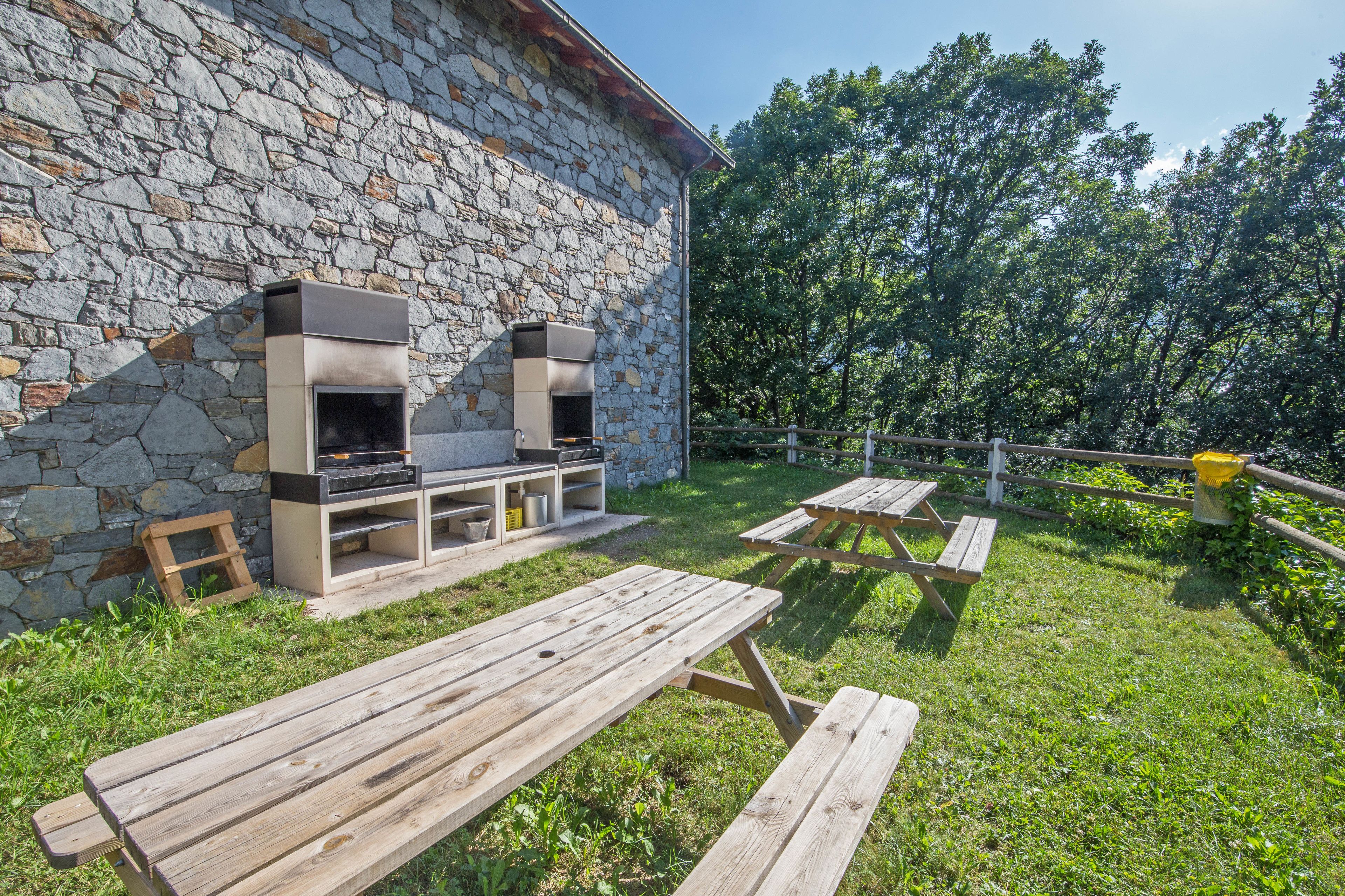 Un edificio de piedra con chimenea de piedra y zona de barbacoa. Hay mesas de picnic y un banco de madera.