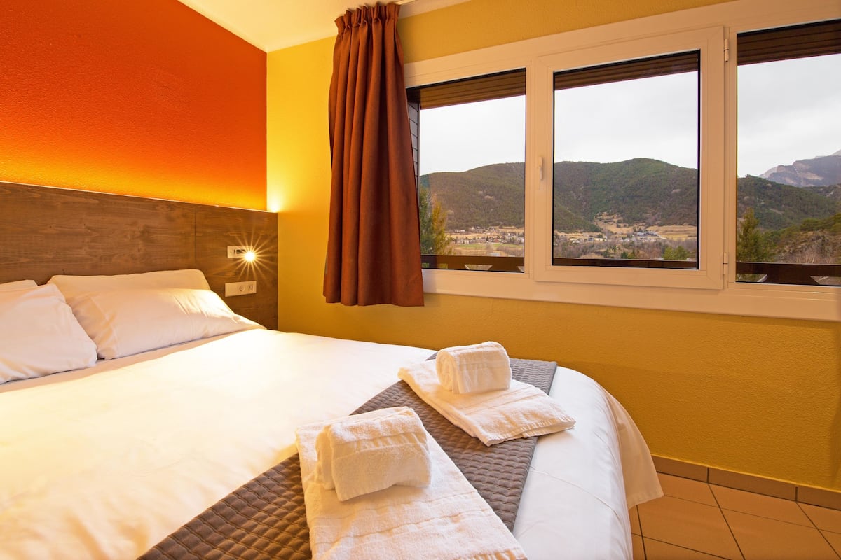 Quarto de hotel com uma cama grande, vista para o campo pela janela e cabeceira com luminária.