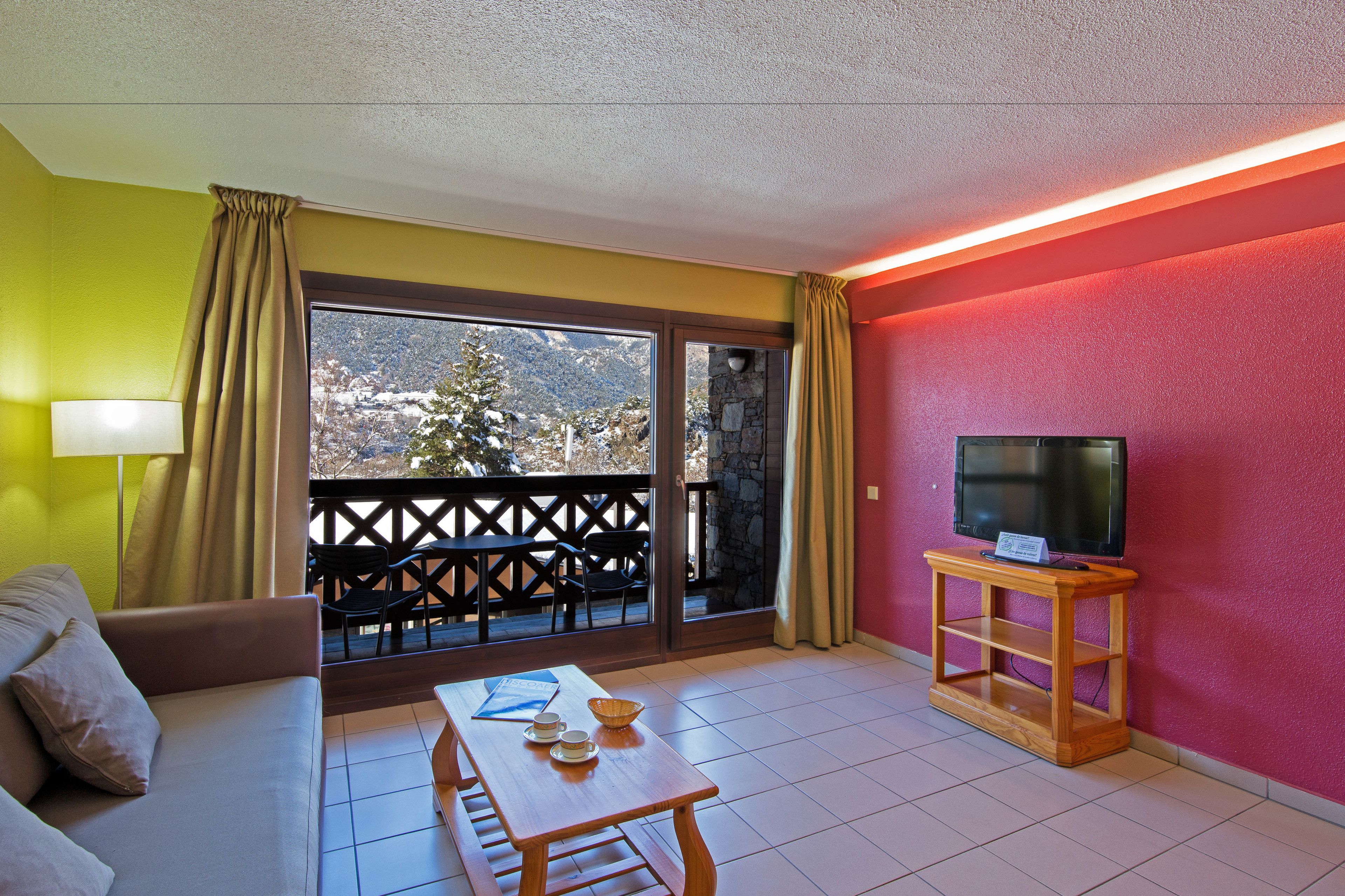A room with a sofa, a table with cups and a bowl, a TV on a wooden stand, and a balcony with a view of snow-covered mountains.