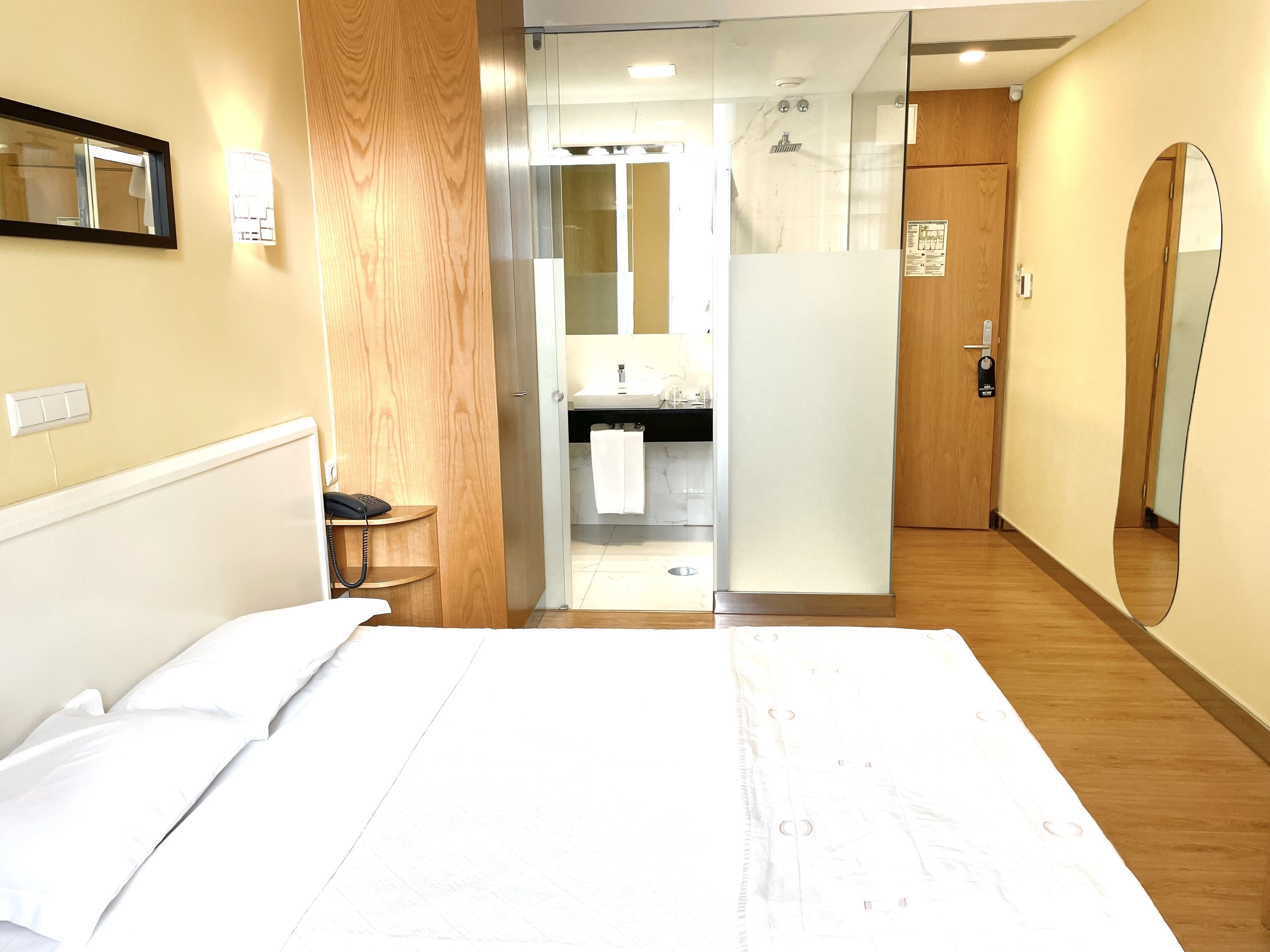 A hotel room with a bed, a wooden side table, a bathroom with a shower, and a mirror.