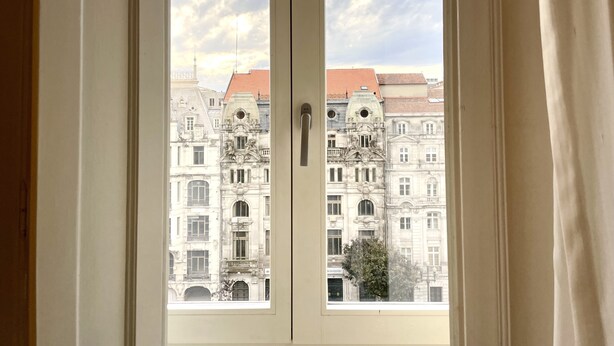 Vista a través de una ventana de un edificio de arquitectura ornamentada, con múltiples ventanas y un tejado de tejas rojas.