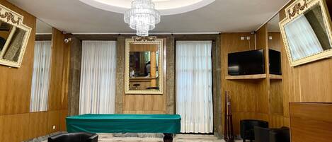 Une pièce avec une table de billard, des fauteuils en cuir noir, un lustre et des boiseries.