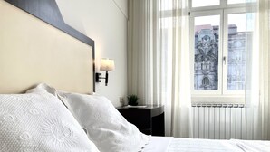 Habitación de hotel con una cama grande, una mesita de noche con lámpara, vista a un edificio por la ventana y un radiador.