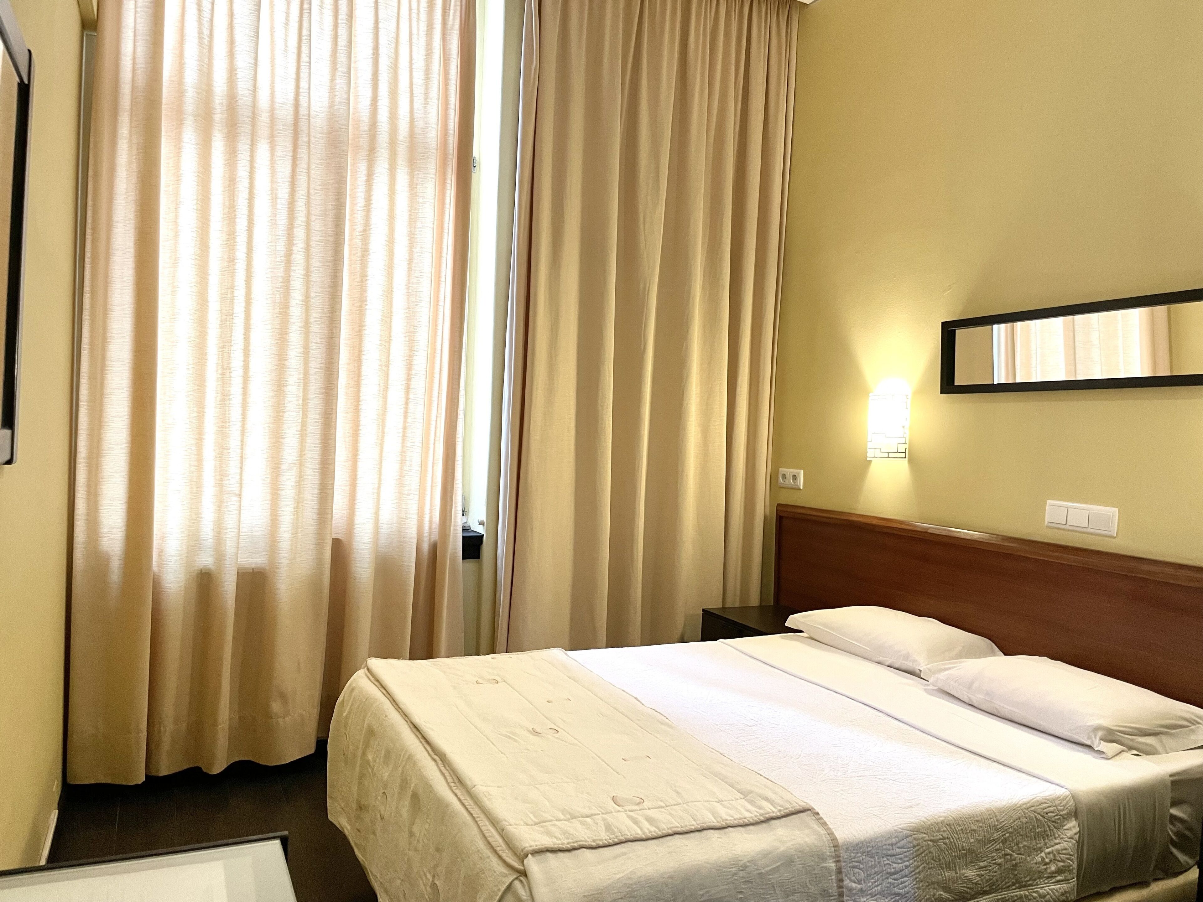 A hotel room with a bed, curtains, a nightstand, a wall-mounted light, and a mirror.