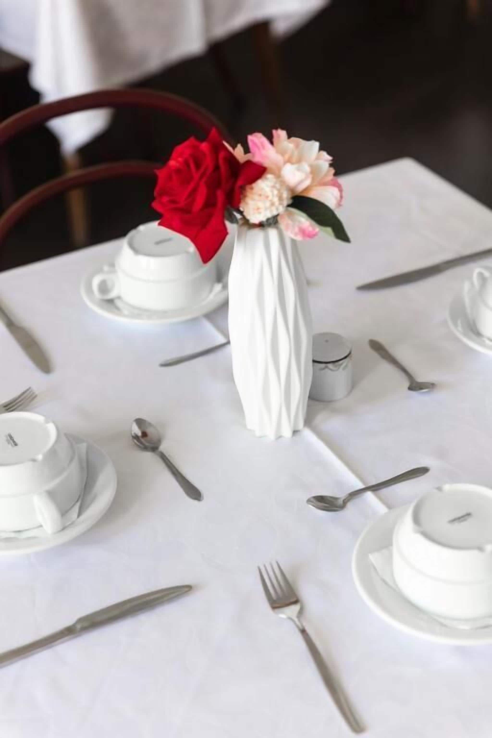A table set with white plates, bowls, and silverware, centered with a white vase containing pink and red flowers.