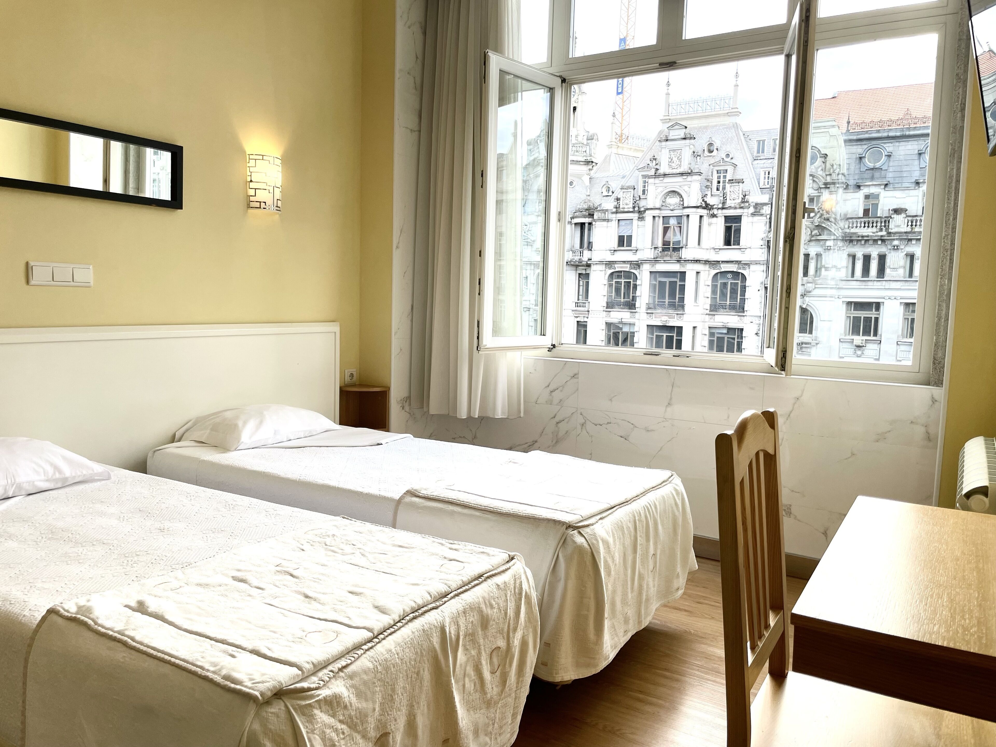 A hotel room with two beds, a wooden chair, a table, a window with a view of historic buildings, and a wall-mounted light fixture.