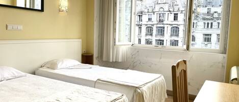 A hotel room with two beds, a wooden chair, a table, a window with a view of historic buildings, and a wall-mounted light fixture.