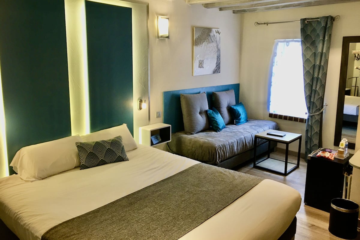 A modern hotel room with a large bed, a sofa, a small table, and a window with curtains.