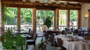 A restaurant with brick walls, large windows, and indoor plants. Tables are set with white tablecloths and have various food items and condiments. - Oasis (Villaines-la-Juhel)