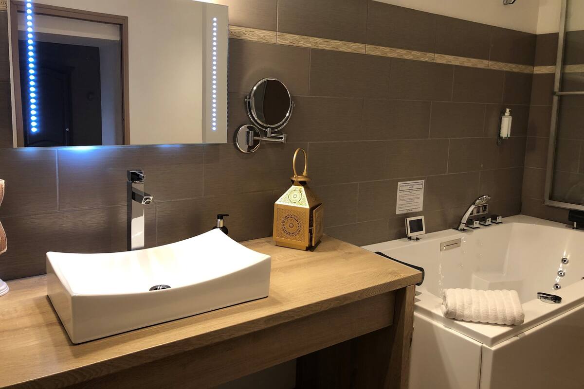 A bathroom with a white sink, a round mirror, a handheld showerhead, a handheld lamp, and a bathtub with a towel.
