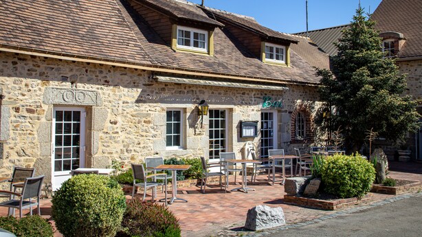 A stone building with a sign reading "l'Oasis." It has outdoor seating with tables and chairs, and a small garden area with bushes and a tree. - Oasis (Villaines-la-Juhel)
