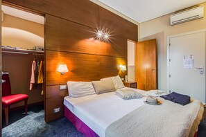 A hotel room with a wooden headboard, a bed with white linens, two bedside lamps, a red chair, a suitcase, and a hat on the bed.
