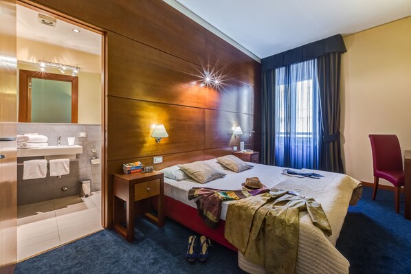 A hotel room with a wooden headboard, a bed with a red bedspread, two bedside lamps, a mirror, a vanity with a sink and towel rack, and a red chair.