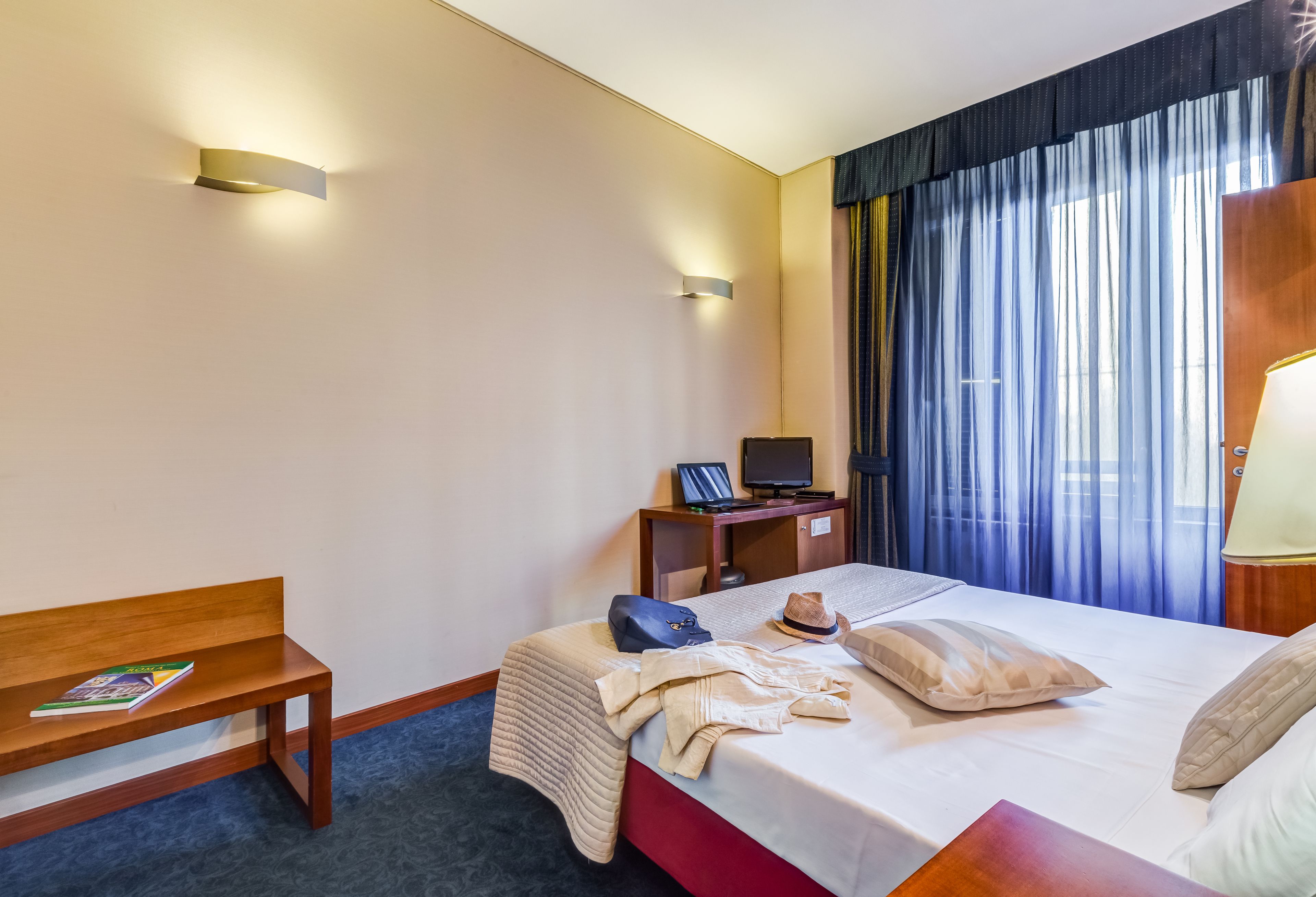 A hotel room with a bed, a desk, a chair, a lamp, and a window with curtains.