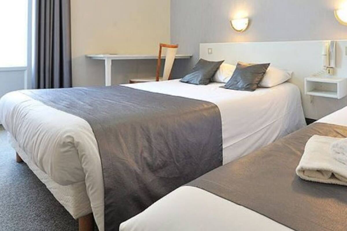 A hotel room with two beds, each with a gray and white bedspread, a nightstand with a lamp, and a view of the outside through a window with curtains.