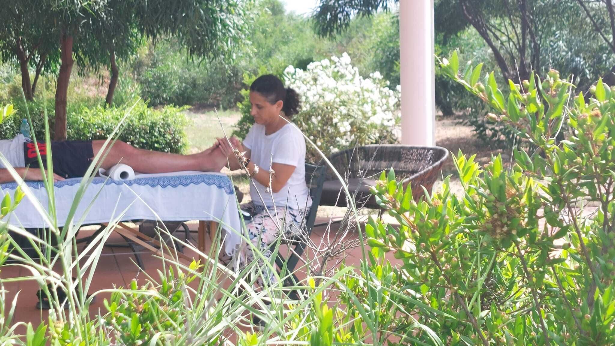Une personne est assise à une table en plein air, recouverte d’une nappe blanche, et est entourée de verdure et d’une structure blanche.
