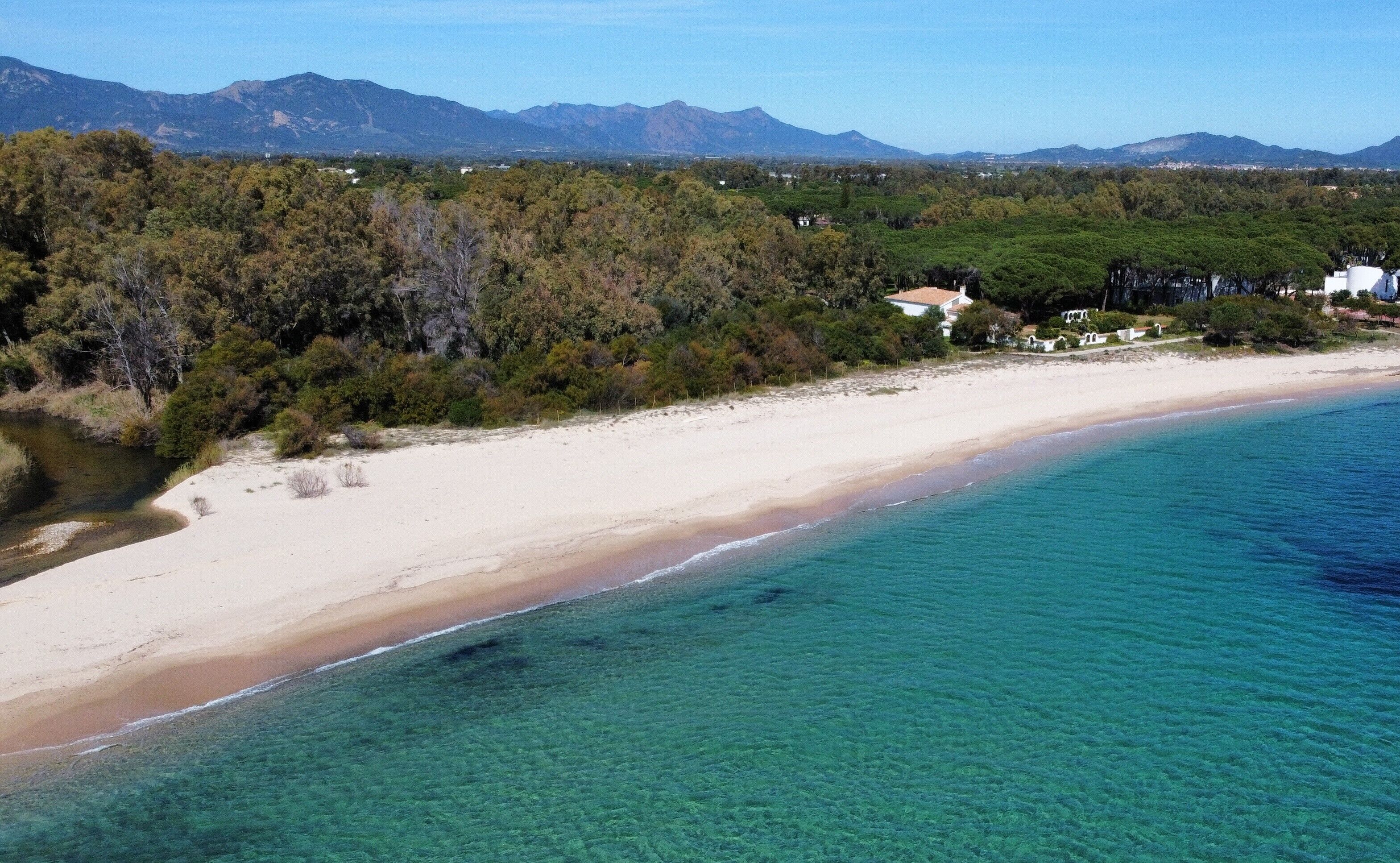 A coastal landscape with a sandy beach, clear turquoise waters, and a backdrop of mountains and dense greenery.