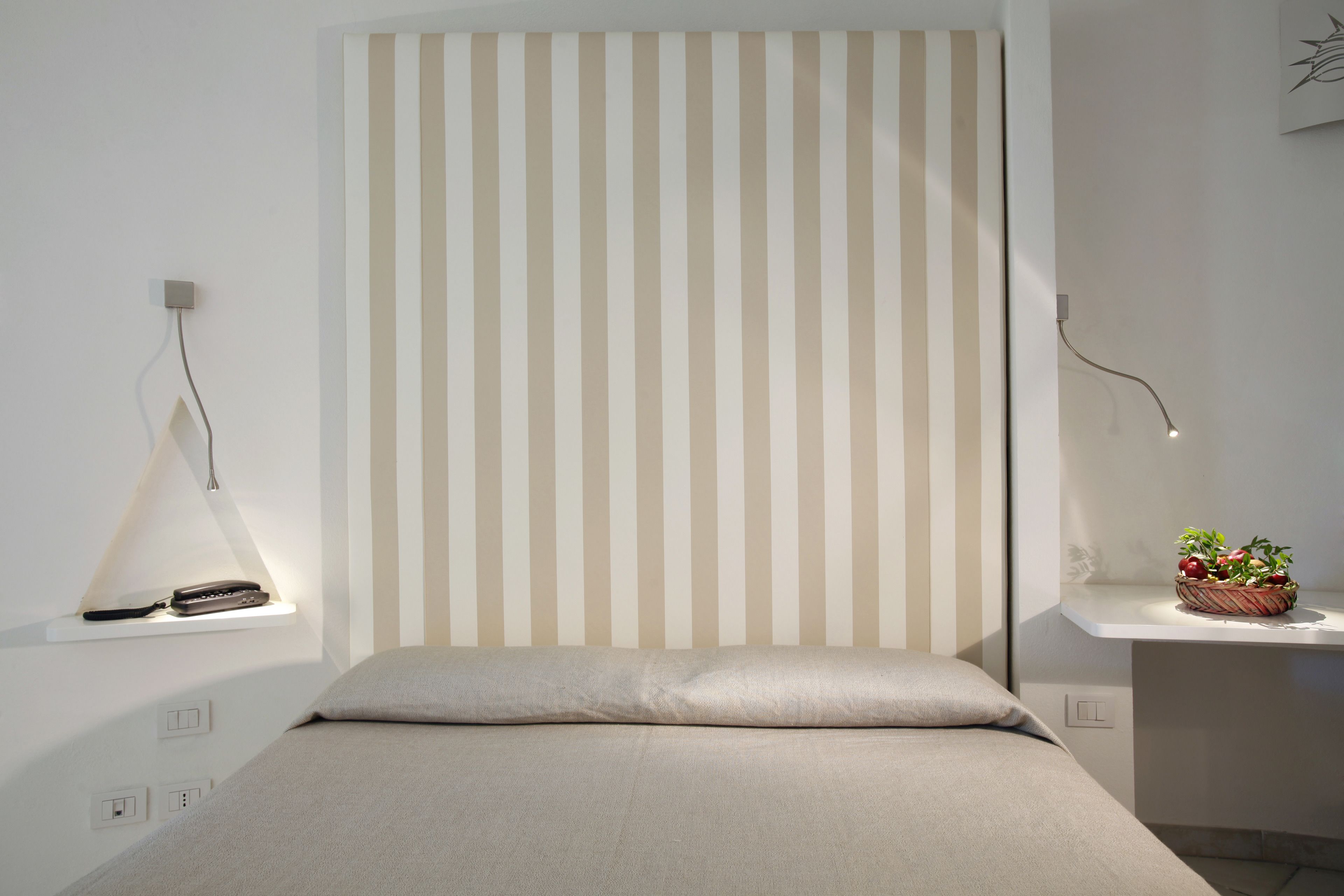 Une chambre à coucher moderne avec une tête de lit à rayures, une table de chevet sur laquelle se trouve un panier de fruits et une lampe suspendue.