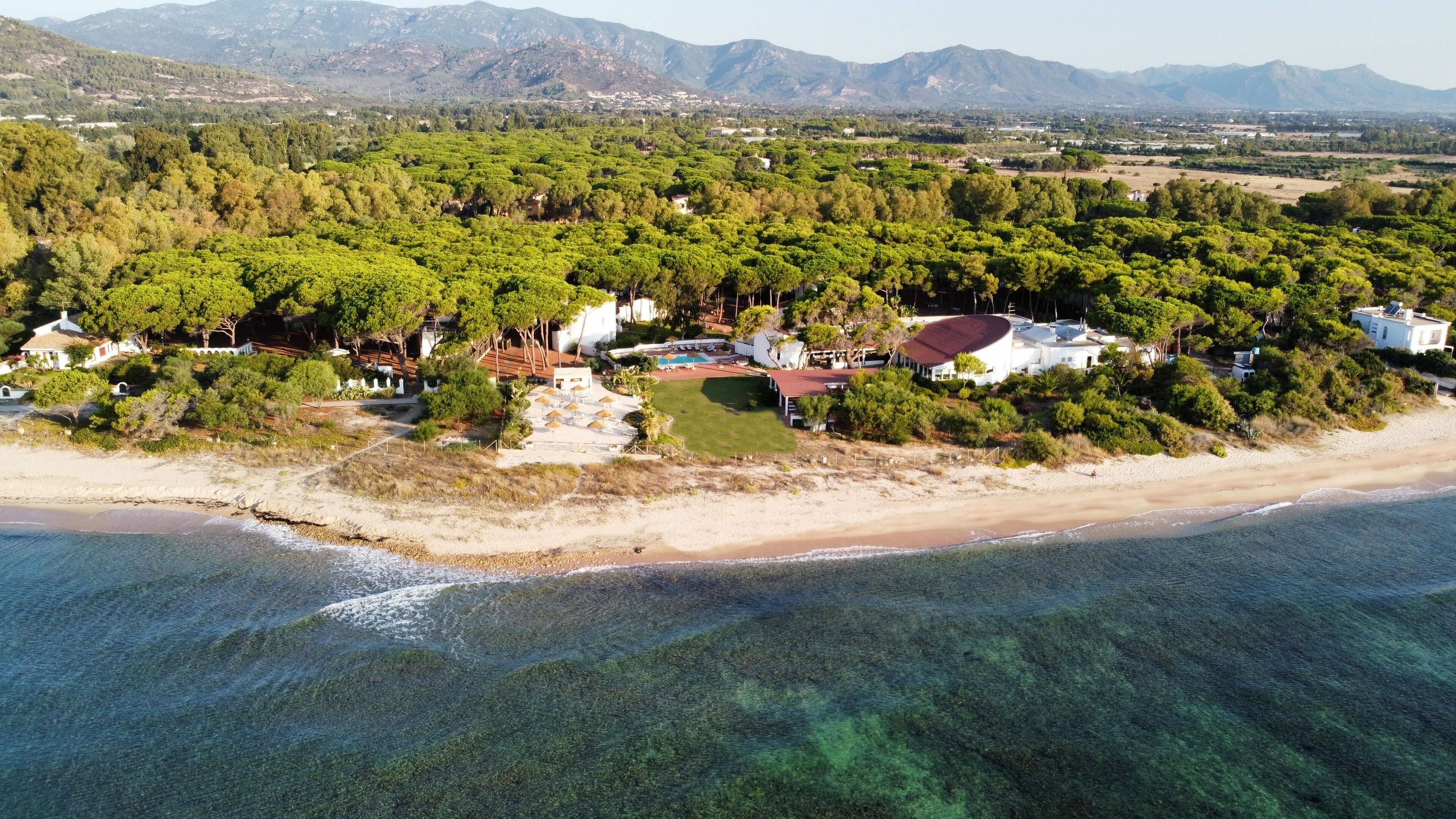 An aerial view of a coastal property with a beach, dense greenery, and a mountainous backdrop.