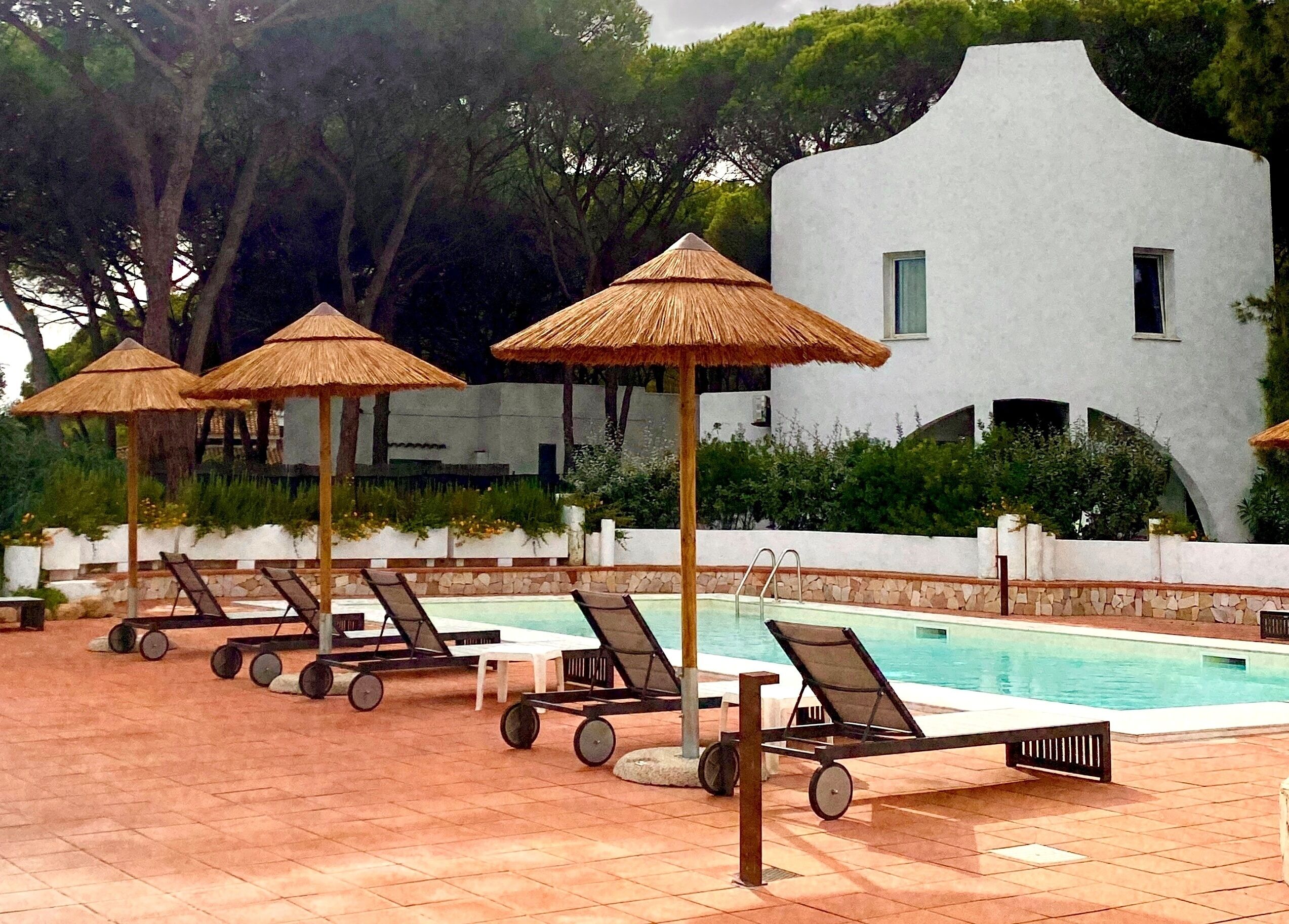 A pool area with lounge chairs and thatched umbrellas, a pool, and a white building with arched windows.