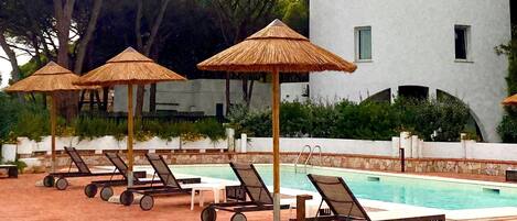 A pool area with lounge chairs and thatched umbrellas, a pool, and a white building with arched windows.