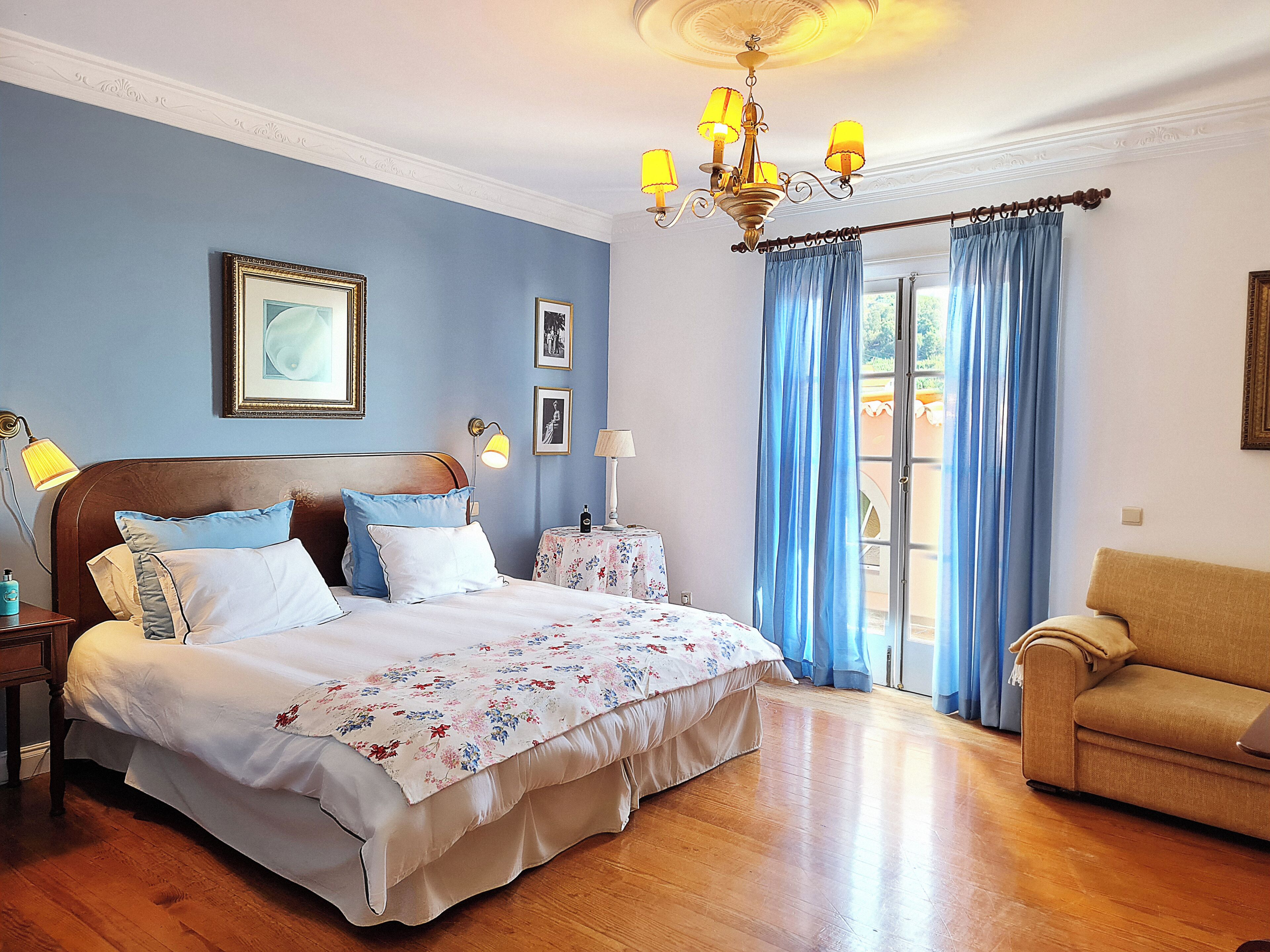 A bedroom with a bed, bedside tables, a chandelier, a sofa, and a view of the outdoors.