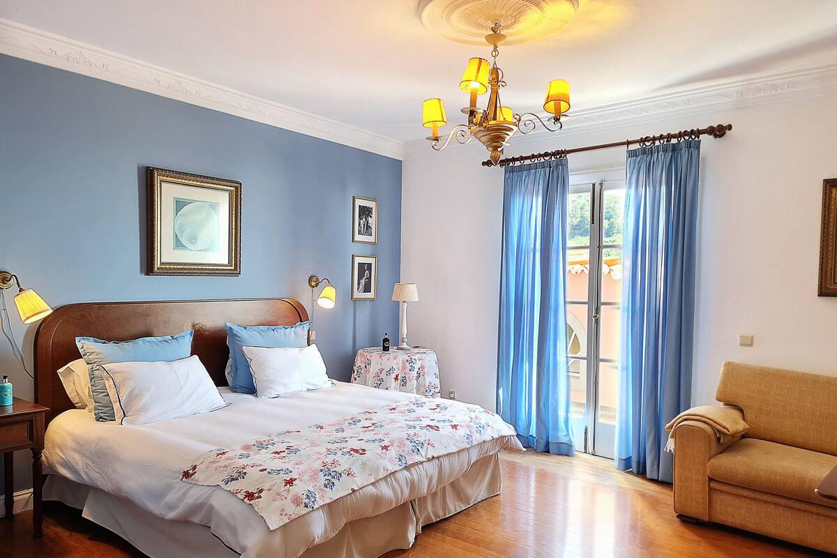 Een slaapkamer met een bed, nachtkastjes, een kroonluchter, een bank en uitzicht op de omgeving.