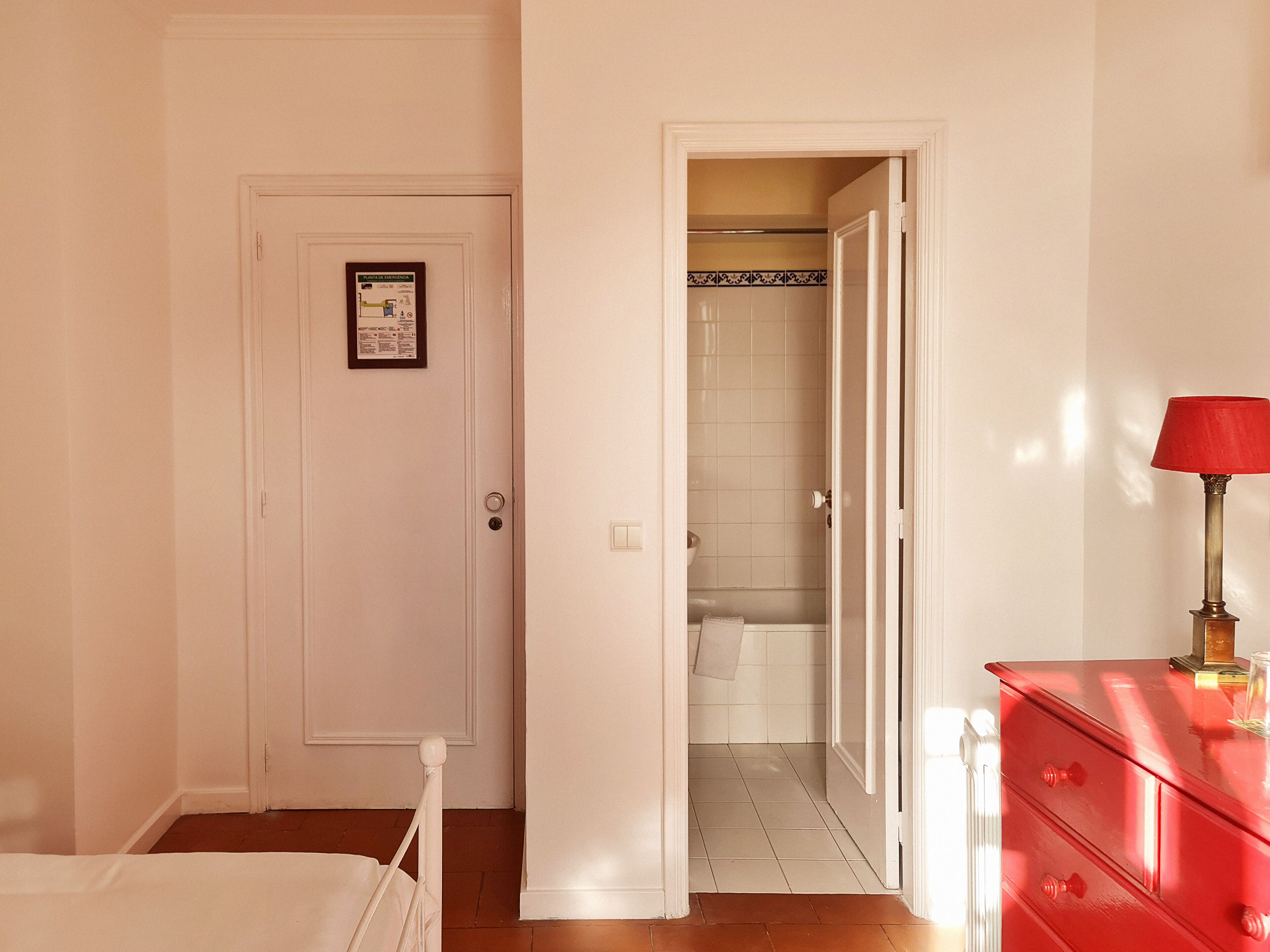 Habitación de hotel con una cama, un aparador rojo, una puerta blanca y un acceso abierto a un baño con ducha e inodoro.