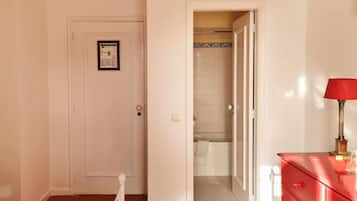Habitación de hotel con una cama, un aparador rojo, una puerta blanca y un acceso abierto a un baño con ducha e inodoro.