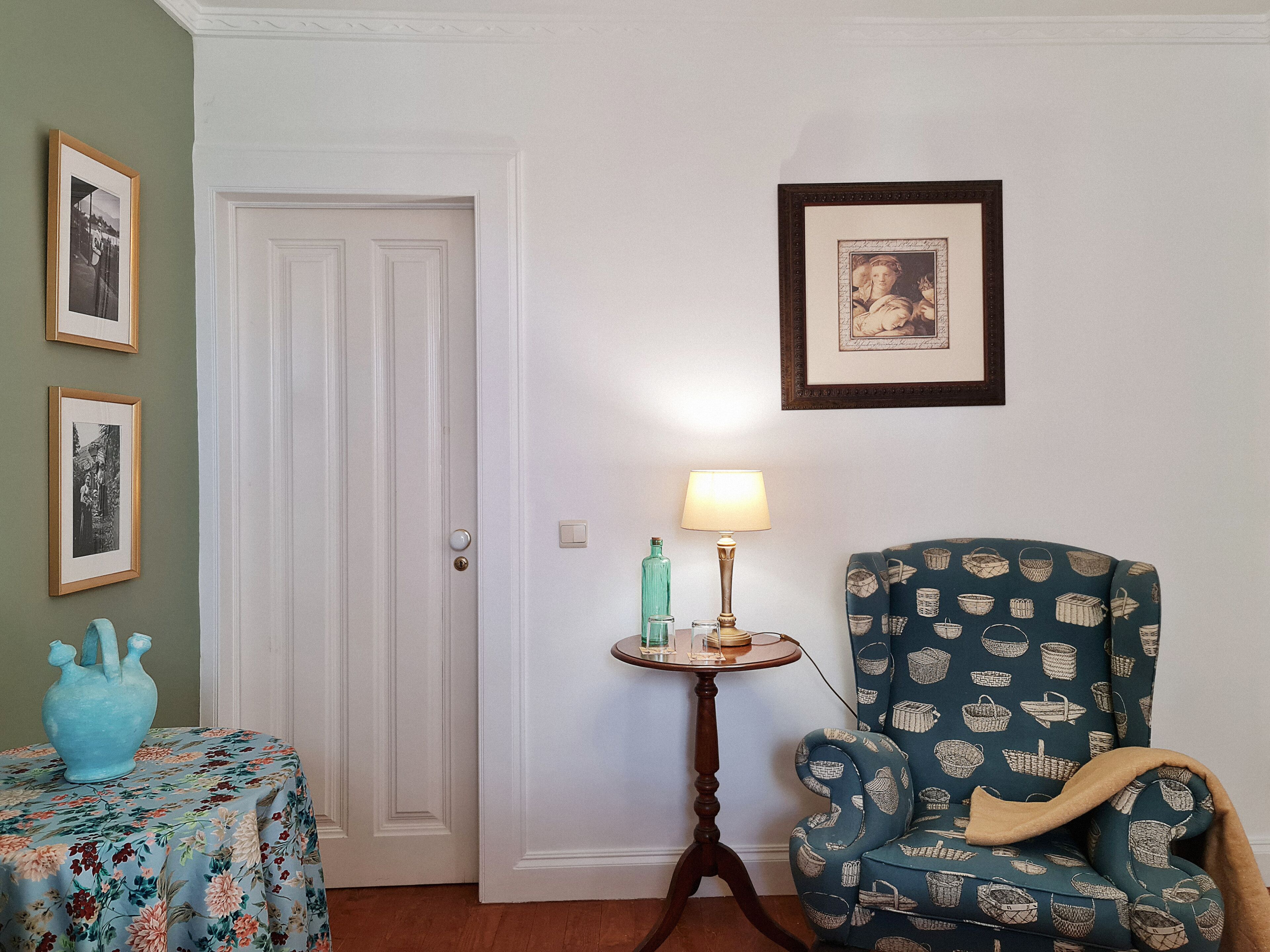 Una habitación con una puerta, una mesita con una lámpara, un sillón estampado y dos cuadros en la pared.