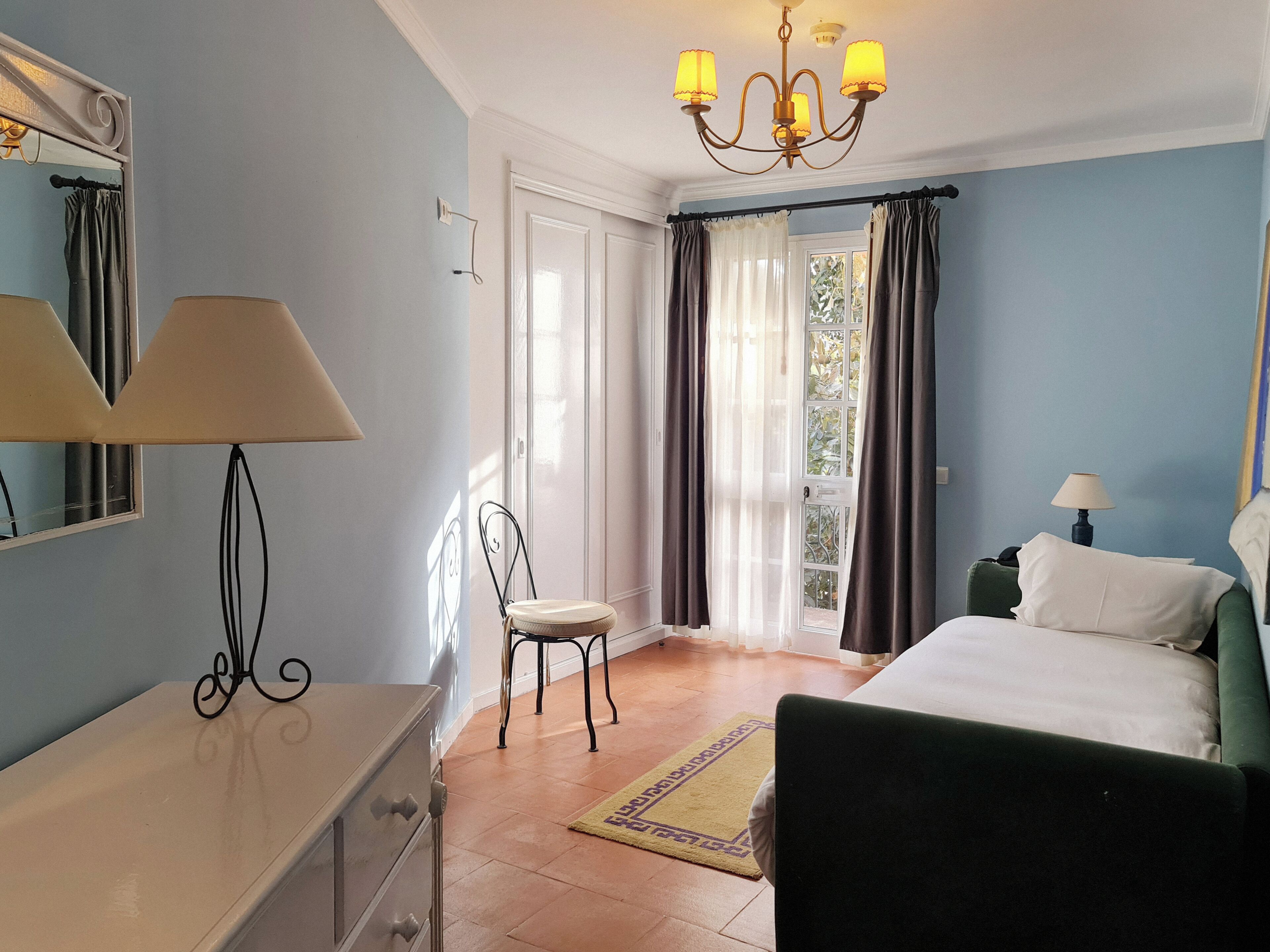 A bedroom with a bed, a chair, a dresser, a lamp, and a chandelier.