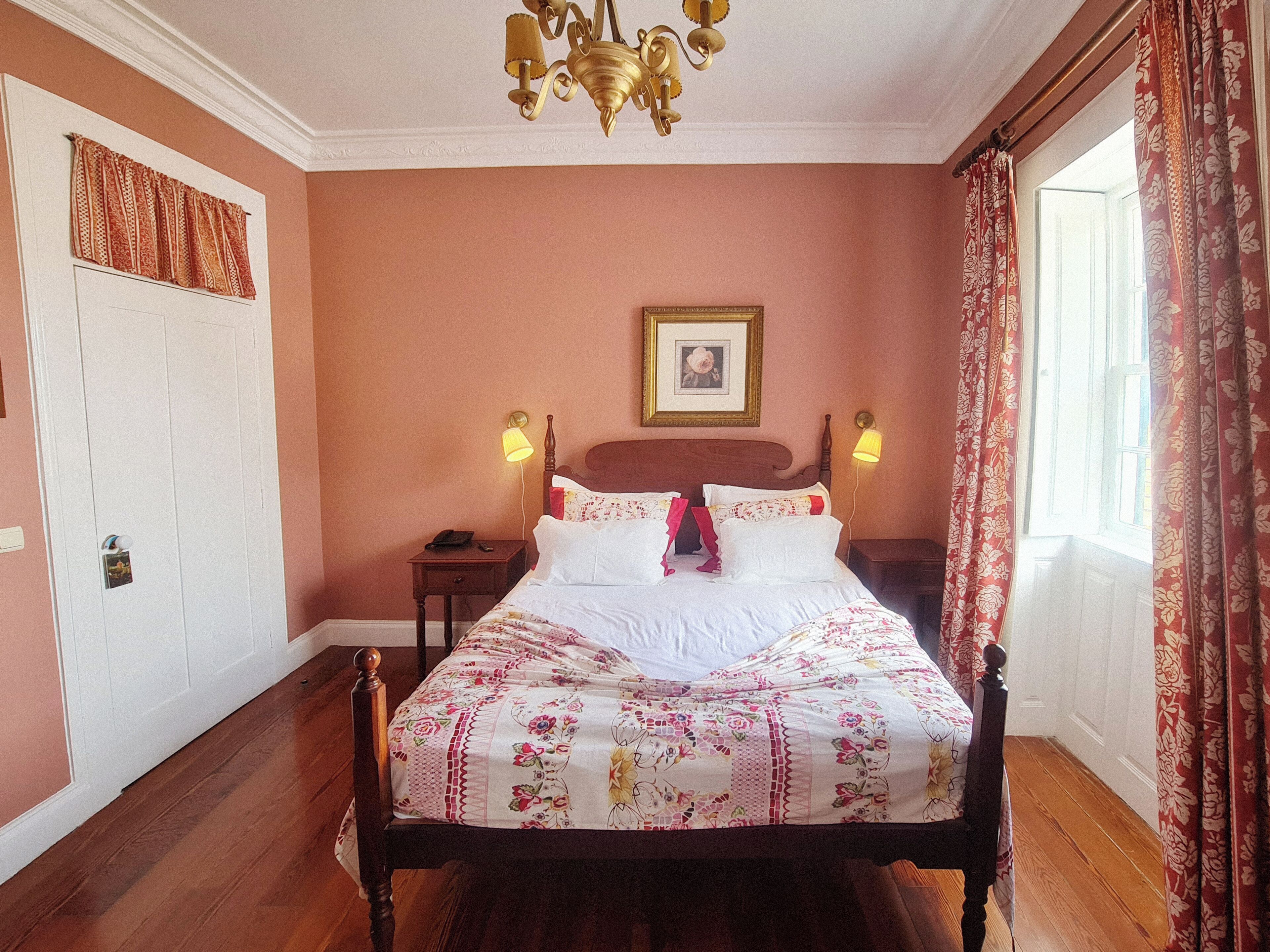 Un dormitorio con cama de madera, una mesita de noche, una lámpara de pared, un cuadro enmarcado y una ventana con cortinas.