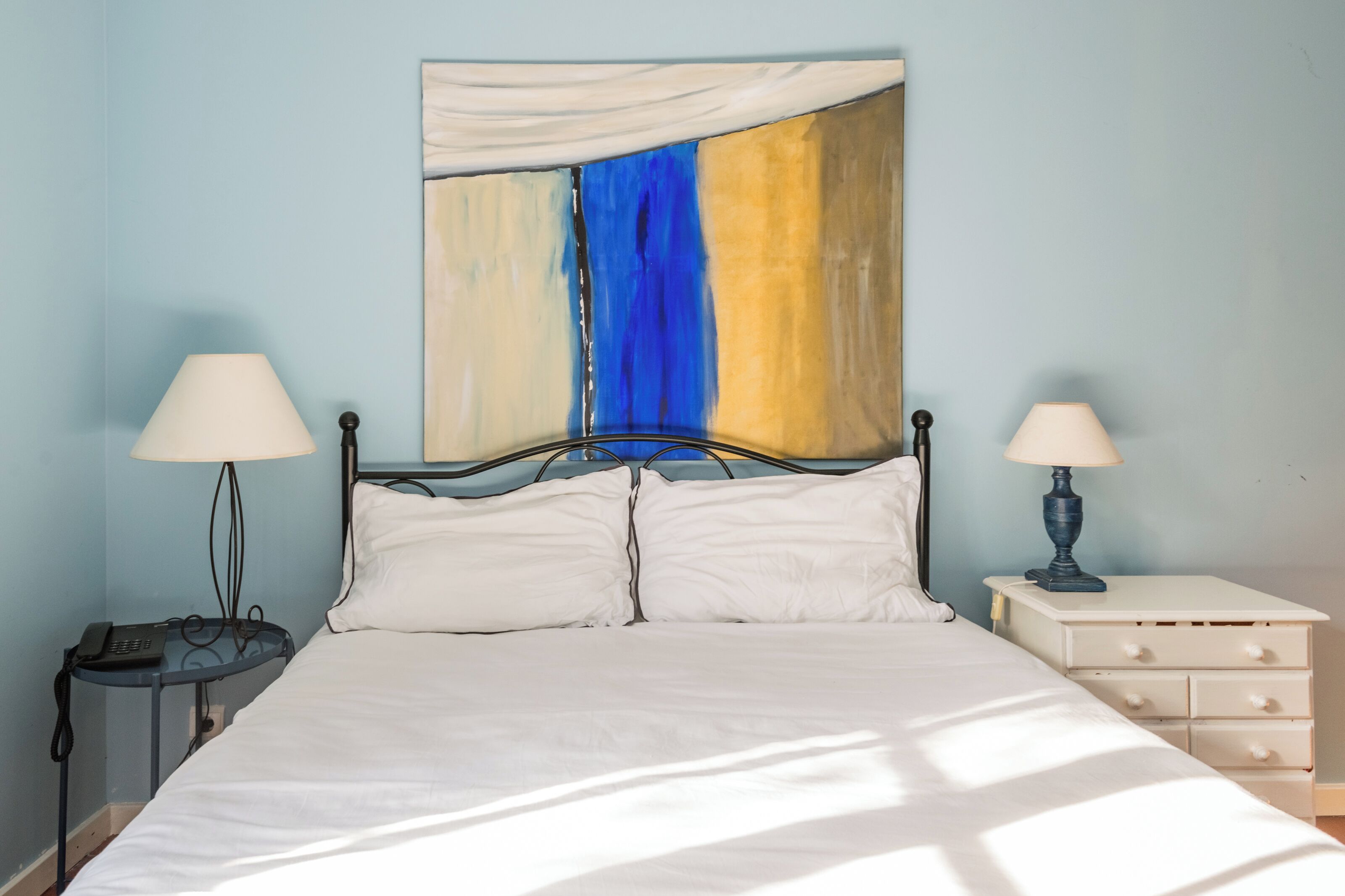A bed with white linens, two bedside tables with lamps, a wall art piece, and a nightstand with drawers.