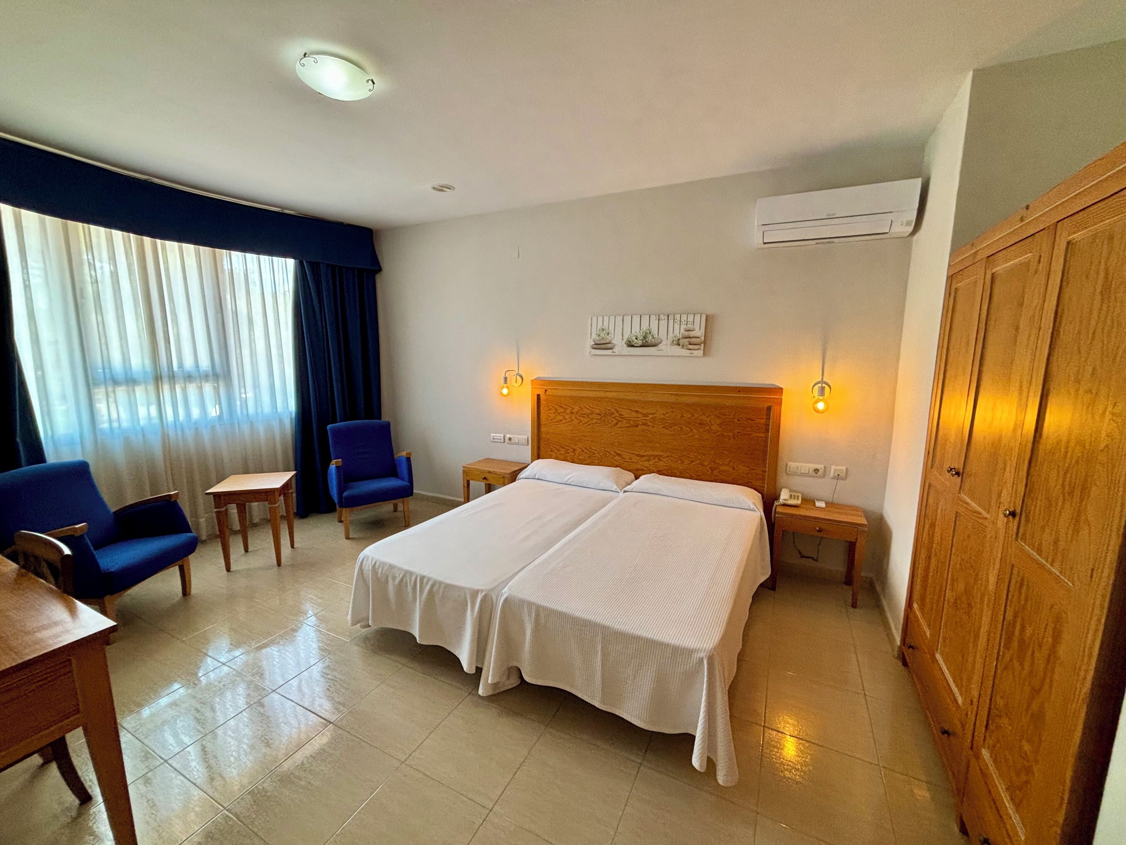 A hotel room with a double bed, two chairs, a small table, a nightstand, and a wardrobe.
