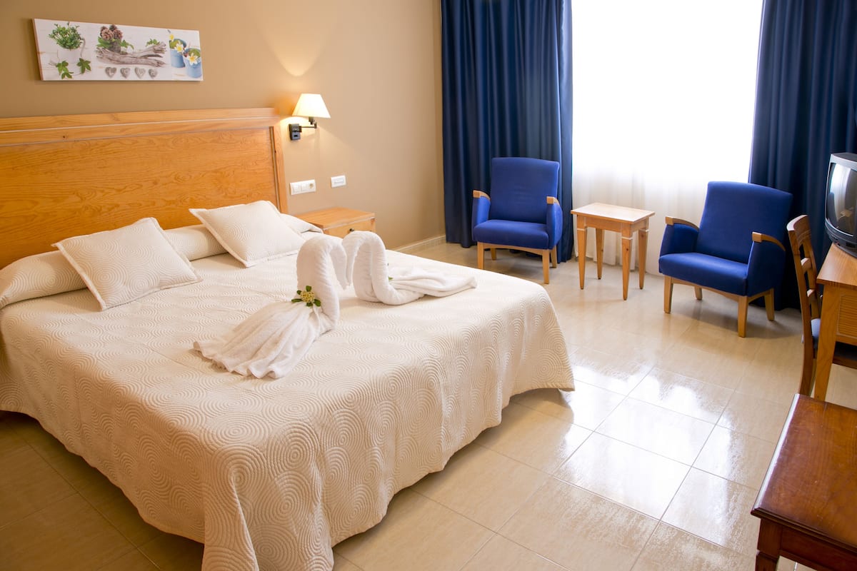 A hotel room with a large bed, two blue armchairs, a small table, a TV, and a lamp.