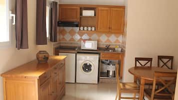 Een kleine keuken met houten kastjes, een wasmachine, een magnetron, en een eethoek met een tafel en stoelen.