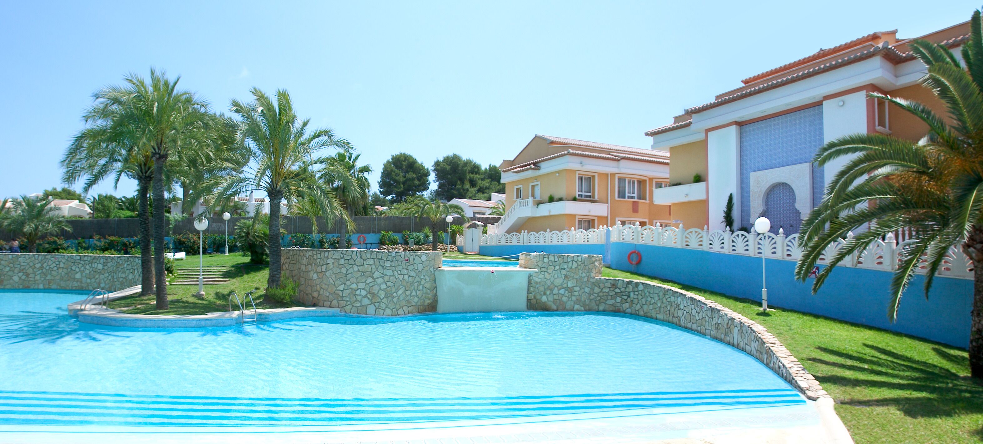 A swimming pool with a stone border, palm trees, and a building with a blue and white facade.