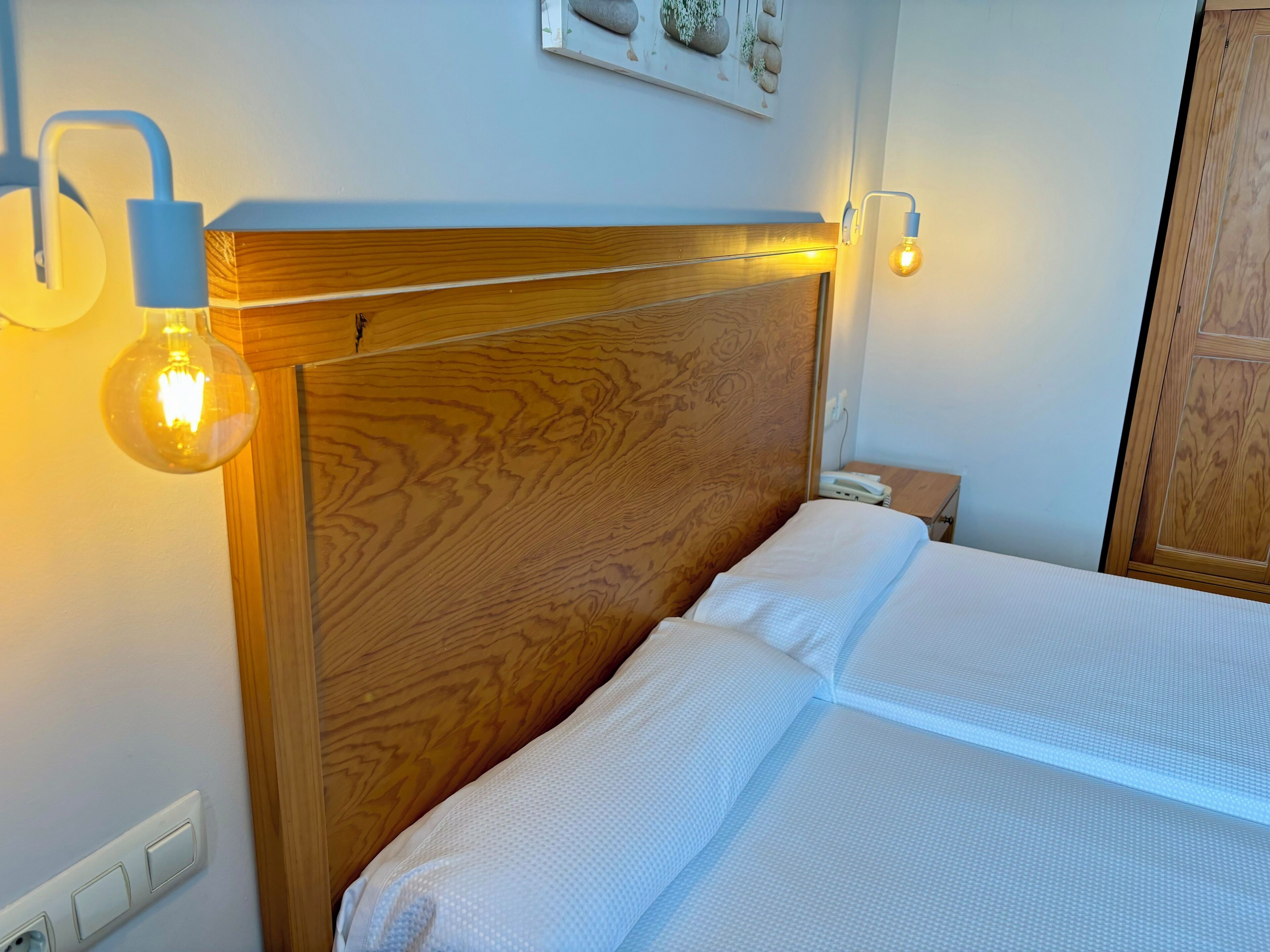A neatly made bed with a wooden headboard, a bedside table with a lamp, and a wall-mounted light fixture.