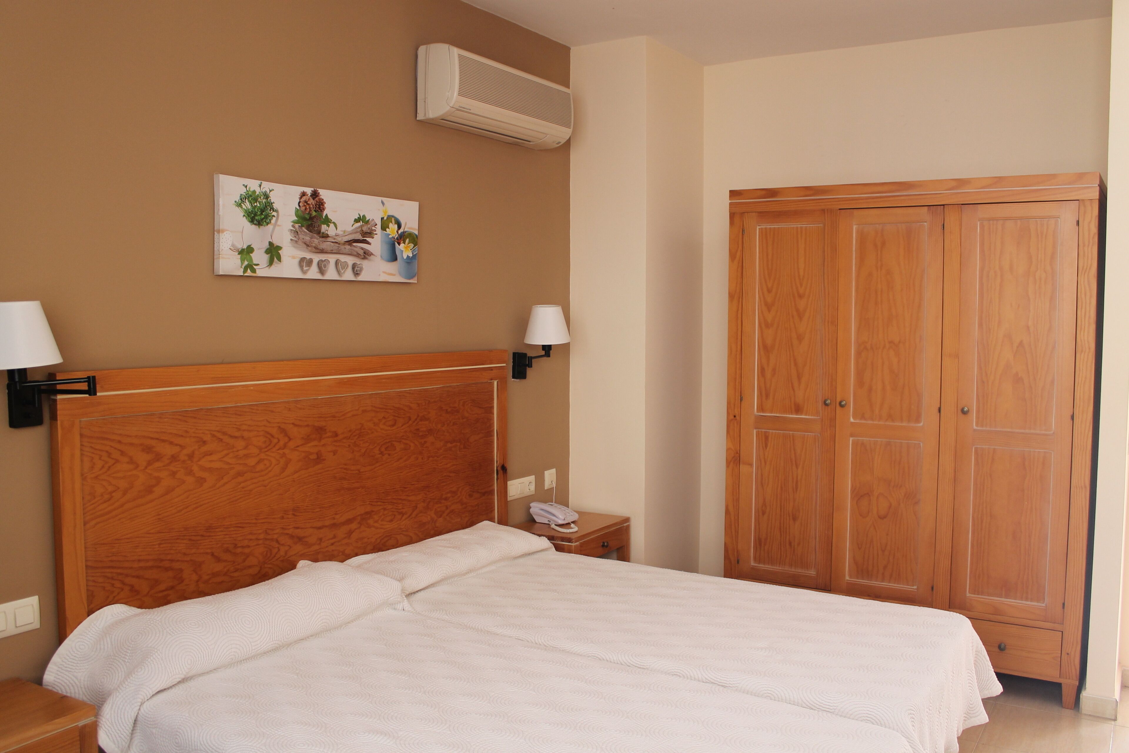 Een slaapkamer met een houten hoofdbord, twee bedlampen, een nachtkastje met een telefoon en een houten garderobe.